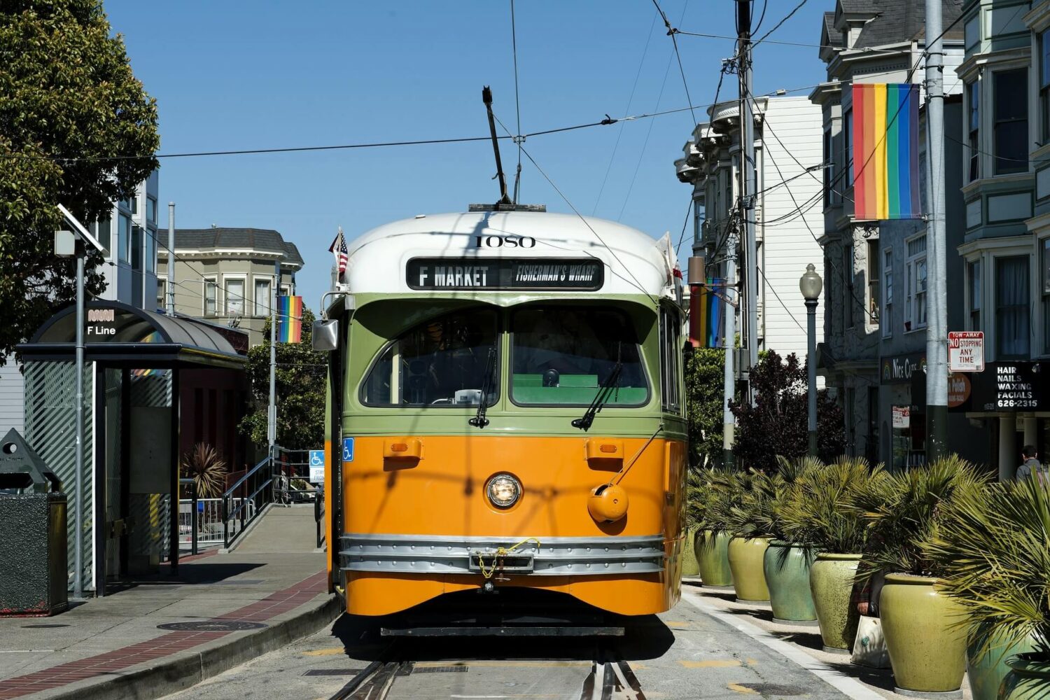 A yellow and green F Line Streetcar in San Francisco with rainbow flags hanging from buildings on either side