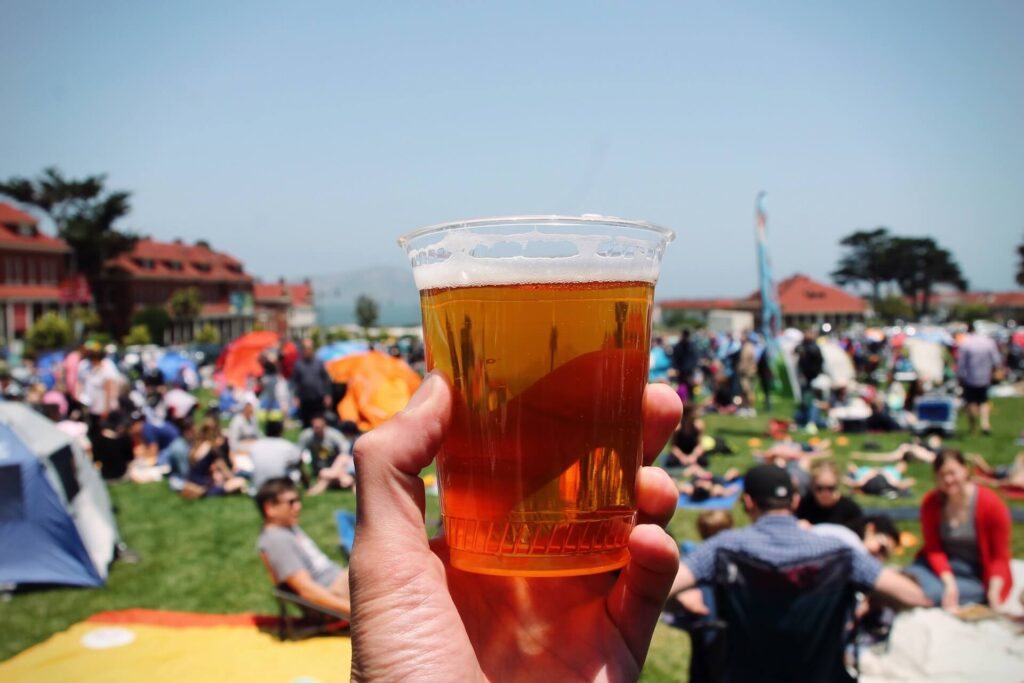 A beer being held up in front of lots of people sitting on the grass on a sunny day in San Francisco